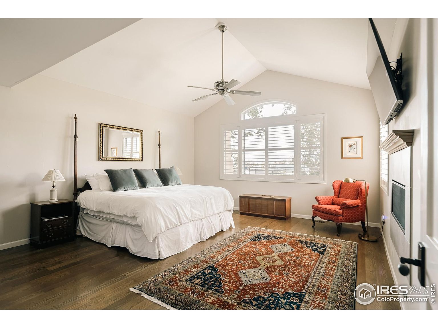 This is a primary bedroom featuring a four-poster bed with white linens and gray pillows, a dark wood nightstand with a lamp, and a large area rug. The room has vaulted ceilings, a window with shutters, and a fireplace, creating a cozy and inviting atmosphere. The flooring is hardwood, and the walls are painted in a neutral tone.