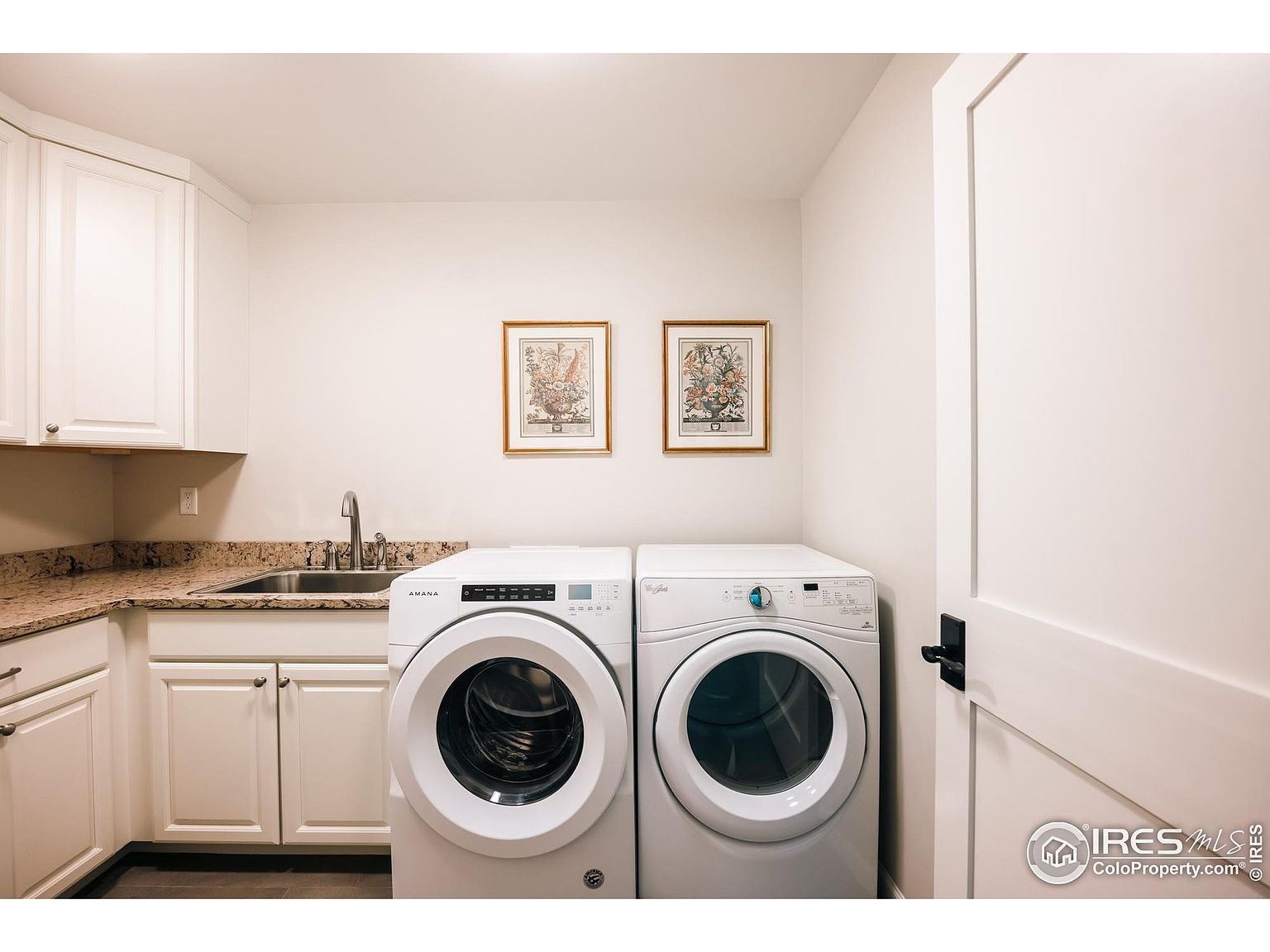 This is an interior shot of a laundry room featuring white cabinetry with granite countertops, a stainless steel sink, and an Amana washing machine paired with a Whirlpool dryer. Two framed botanical prints hang above the appliances, adding a touch of decor to the functional space. A white door with a black handle is visible on the right side of the frame.