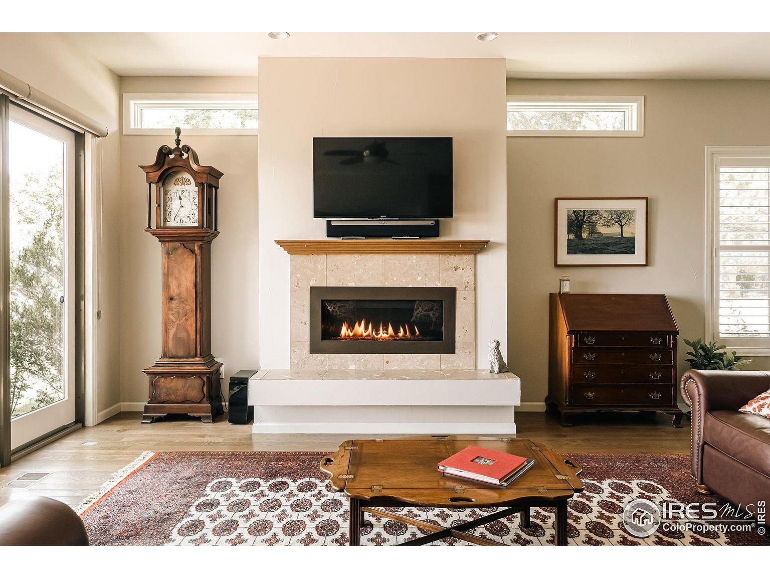 This is an interior shot of a living room featuring a fireplace with a mounted television above it. The room is decorated with a grandfather clock, a wooden desk, and a patterned rug. Natural light streams in through the windows and sliding glass door, creating a warm and inviting atmosphere.