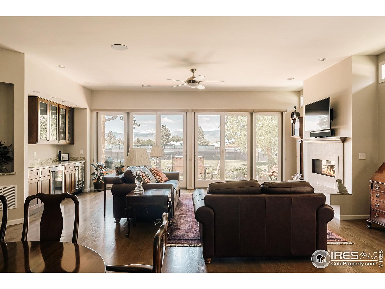 This is an interior shot of a living room featuring hardwood floors, a fireplace with a mounted television, and a wet bar. Large sliding glass doors provide ample natural light and a view of the backyard. The room is furnished with two leather sofas and a dark wood dining table, creating a warm and inviting atmosphere.