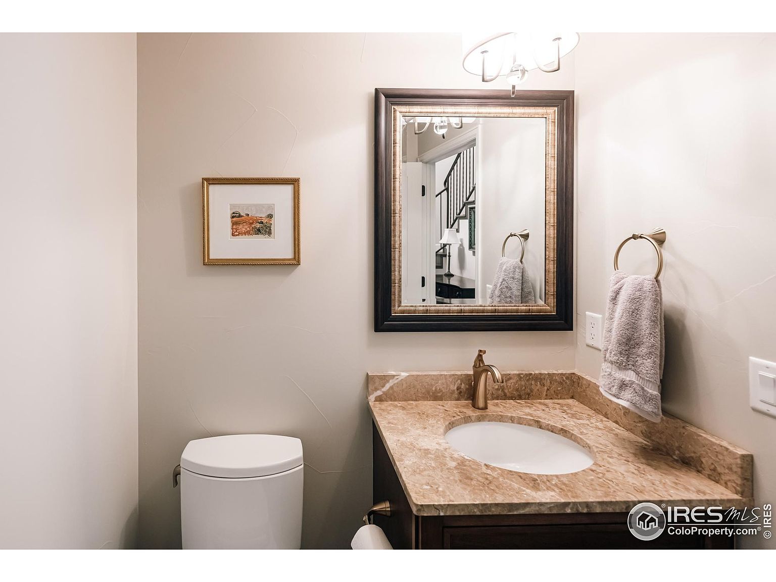 This is a well-lit guest bathroom featuring a vanity with a marble countertop and a dark wood cabinet. A framed mirror hangs above the sink, reflecting a glimpse of the staircase. A framed picture hangs on the wall to the left of the mirror, and a white toilet is visible in the corner.