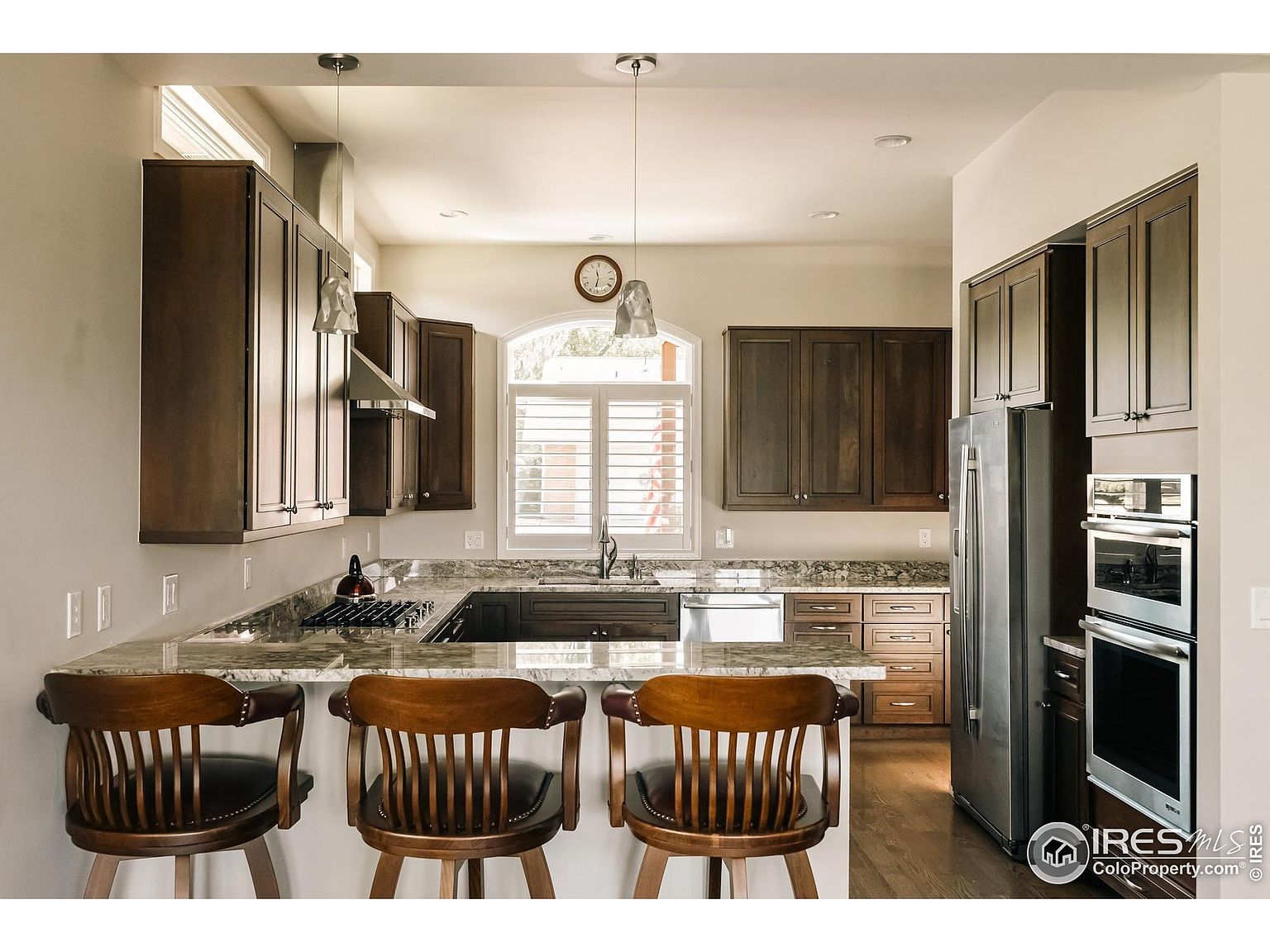 This is a well-lit kitchen featuring dark wood cabinetry and granite countertops. The kitchen includes stainless steel appliances, a breakfast bar with three wooden stools, and a window above the sink. The overall impression is a warm and inviting space, perfect for cooking and entertaining.