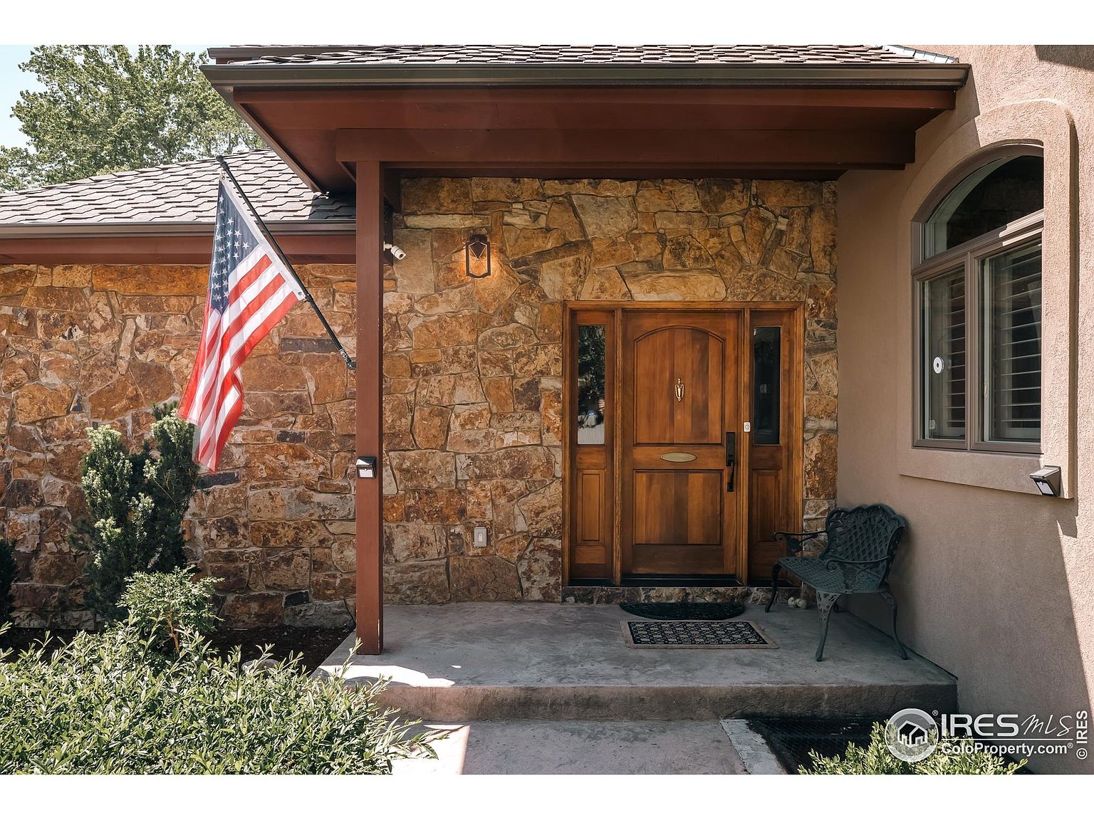 This image showcases the entryway of a home, featuring a stone facade and a wooden front door with sidelights. An American flag is displayed on the left, adding a patriotic touch. A small bench sits to the right of the door, and a window with an arched top is visible on the right side of the entryway. The overall impression is welcoming and well-maintained.