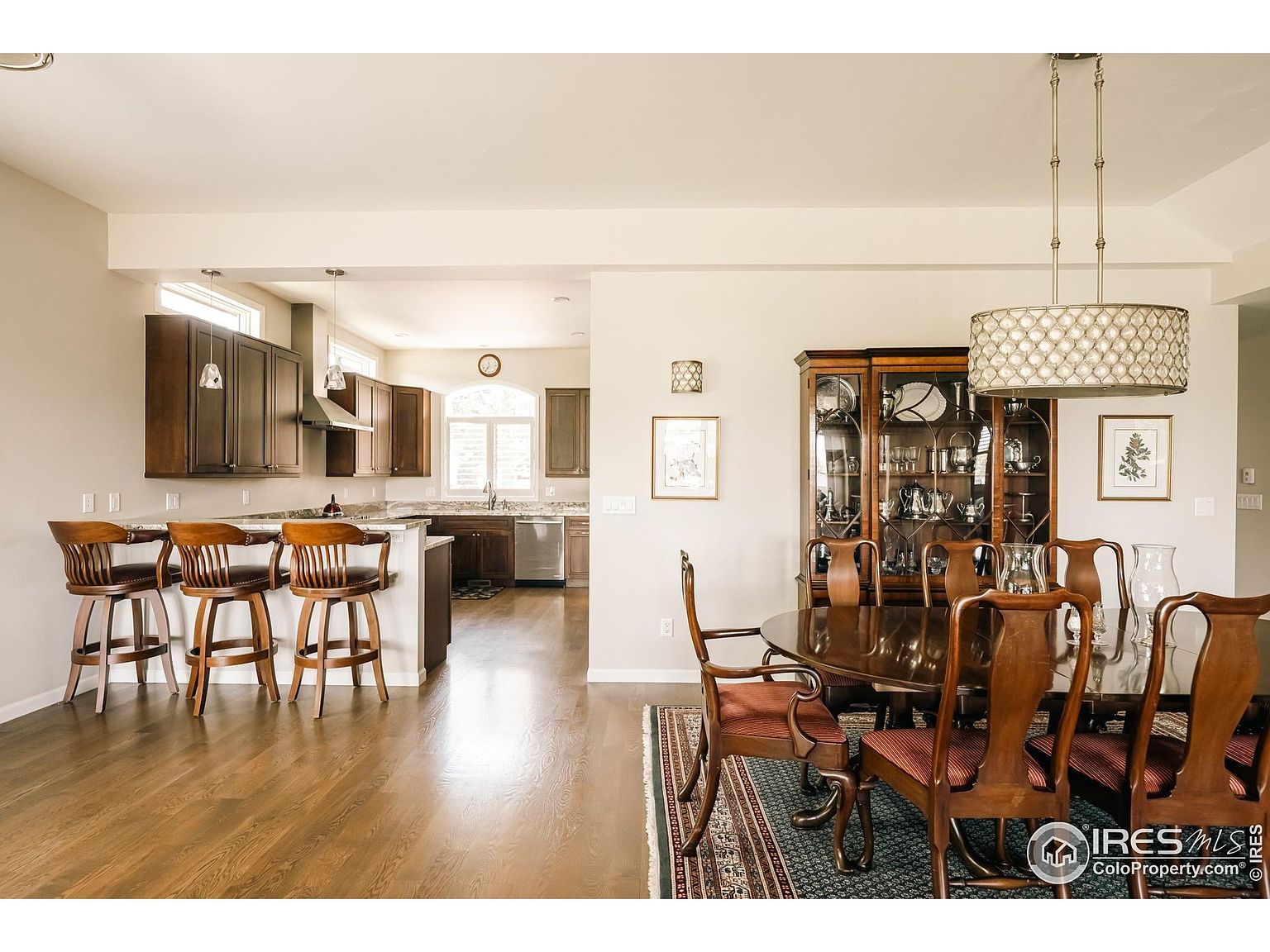 This is an interior shot showcasing a dining room adjacent to a kitchen area. The dining room features a dark wood dining table with chairs, a china cabinet, and a decorative light fixture. The kitchen area includes a breakfast bar with stools and dark wood cabinetry, creating a warm and inviting atmosphere.