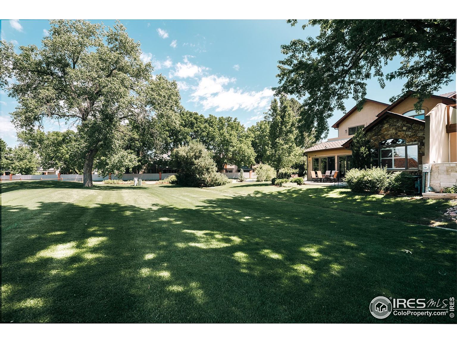 This image showcases a spacious and well-maintained backyard. The lush green lawn is partially shaded by mature trees, creating a serene and inviting atmosphere. In the background, a portion of the house is visible, featuring a stone facade and a sunroom, suggesting a blend of natural and architectural elements.