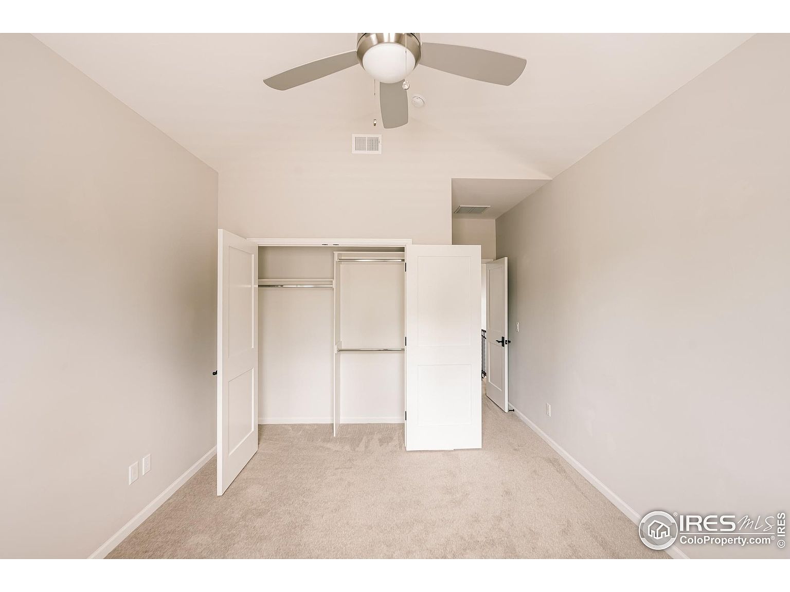 This is a bright and airy bedroom featuring neutral-toned walls and carpet. A closet with white doors is partially open, revealing shelving and hanging rods. A ceiling fan provides air circulation, and a doorway leads to another room, creating a sense of spaciousness.