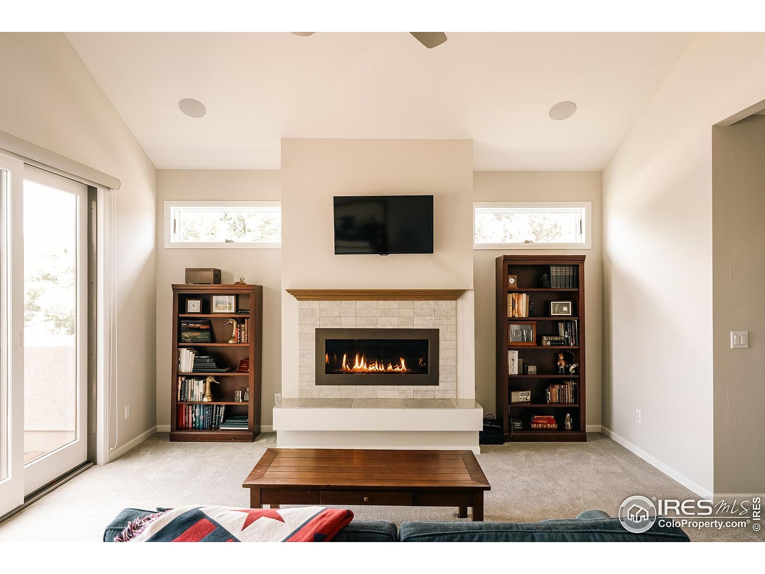 This is an interior shot of a living room featuring a modern fireplace with a television mounted above it. Two wooden bookshelves flank the fireplace, and a wooden coffee table sits in front of a blue sofa. Natural light enters through a sliding glass door on the left, creating a bright and inviting atmosphere.