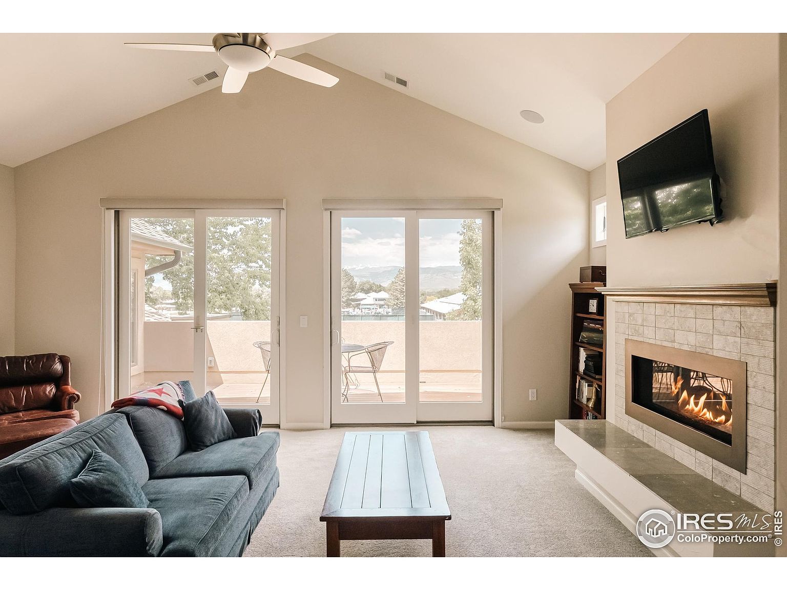This is an interior shot of a living room featuring a vaulted ceiling, a fireplace with a mounted television above, and sliding glass doors leading to a balcony. The room is furnished with a blue sofa and a dark wood coffee table, creating a cozy and inviting atmosphere. The natural light from the balcony doors brightens the space, highlighting the neutral color palette.