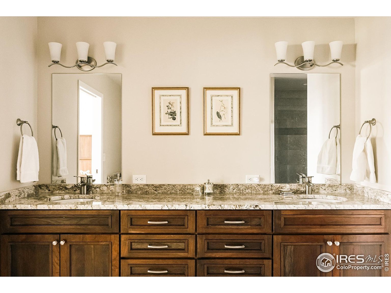 This is a well-lit primary bathroom featuring a double vanity with a granite countertop and dark wood cabinetry. Two framed botanical prints hang above the countertop between the mirrors and light fixtures. The overall impression is clean and elegant, suggesting a comfortable and functional space.