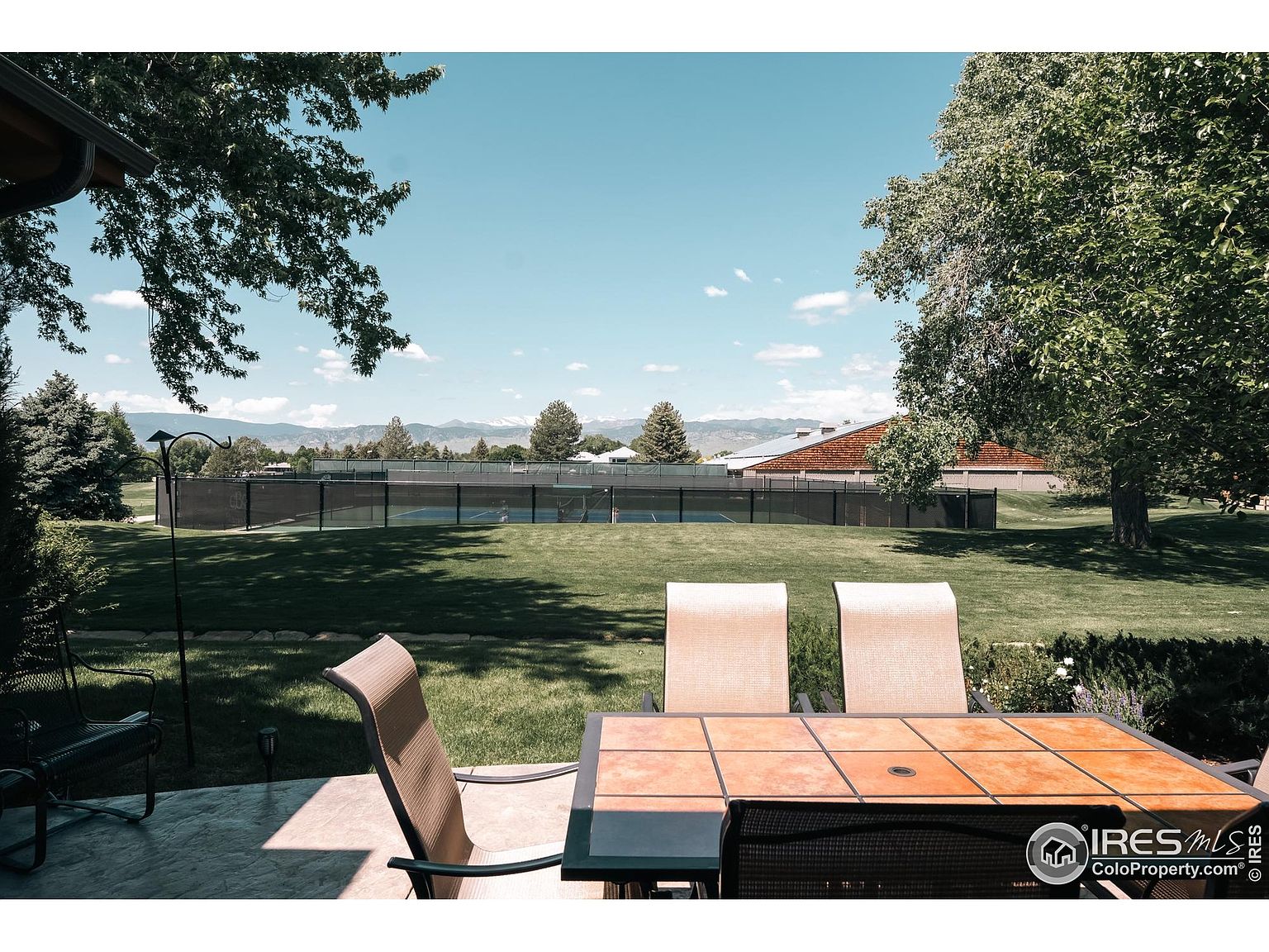 This image showcases a serene outdoor patio setting, featuring a tiled table with chairs, overlooking a well-manicured lawn and a tennis court. Mature trees provide shade and enhance the privacy of the space, while the clear blue sky adds to the inviting atmosphere. The scene suggests a relaxing and private outdoor living area, perfect for entertaining or enjoying quiet moments.