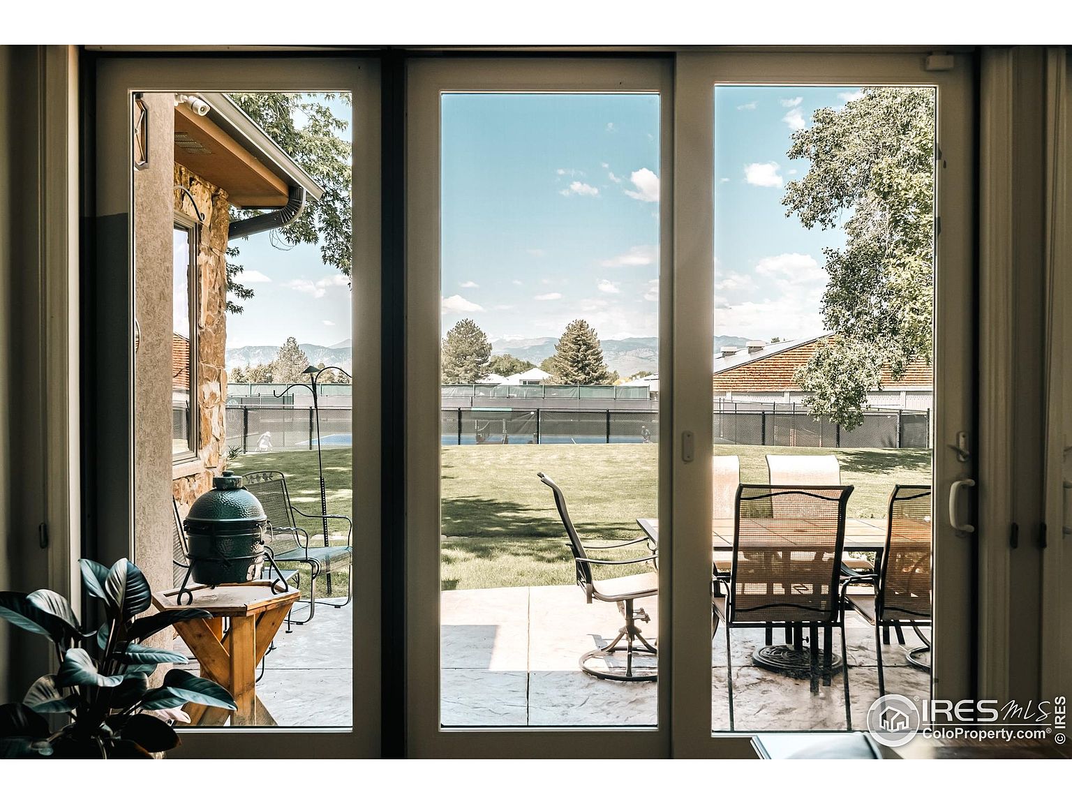 The image showcases an interior view through a large sliding glass door, revealing a well-maintained backyard. Outside, there's a patio area with outdoor dining furniture and a grill, set against a backdrop of lush green lawn and mature trees. The scene evokes a sense of indoor-outdoor living and relaxation.