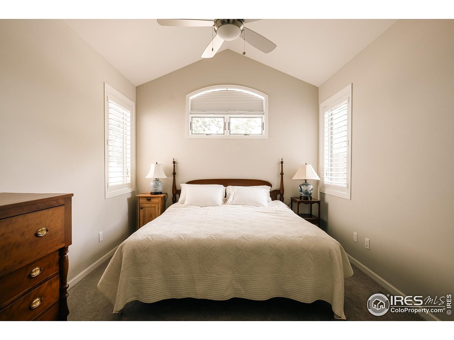 This is a well-lit primary bedroom featuring a queen-sized bed with white bedding, flanked by matching nightstands and lamps. The room has a neutral color palette, plantation shutters on the windows, and a ceiling fan. A wooden dresser is visible on the left side of the frame, adding to the room's traditional style.