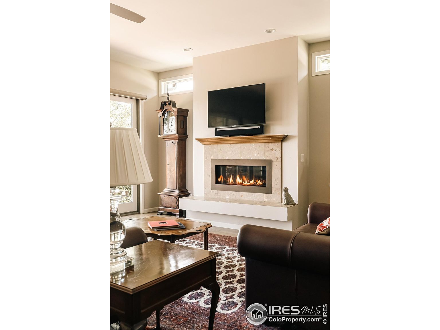 This is an interior shot of a living room featuring a fireplace with a mounted television above it. The room is furnished with a brown leather sofa, wooden coffee tables, and a grandfather clock. The overall style is traditional and cozy, creating a warm and inviting atmosphere.