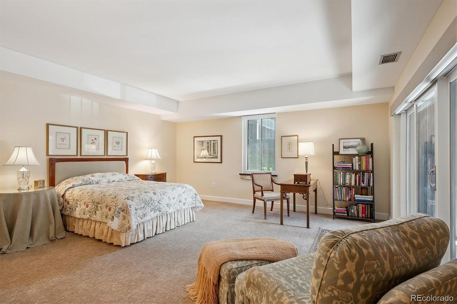 This is a spacious primary bedroom featuring a large bed with floral bedding, complemented by framed artwork above the headboard. The room includes a sitting area with an upholstered chair and ottoman, a desk with a chair, and a bookshelf, creating a comfortable and functional space. Natural light streams in through a window and sliding glass doors, enhancing the room's inviting atmosphere.