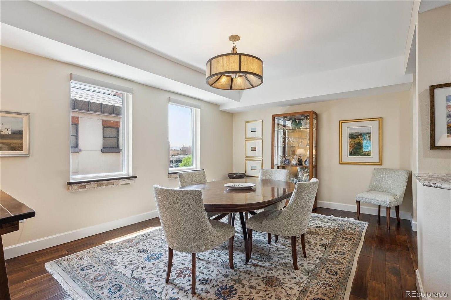 This is a well-lit dining room featuring a round wooden table surrounded by four upholstered chairs. A patterned area rug anchors the space, and a drum-style chandelier hangs above the table. A glass-front cabinet displays decorative items, and artwork adorns the walls, creating a sophisticated and inviting atmosphere.
