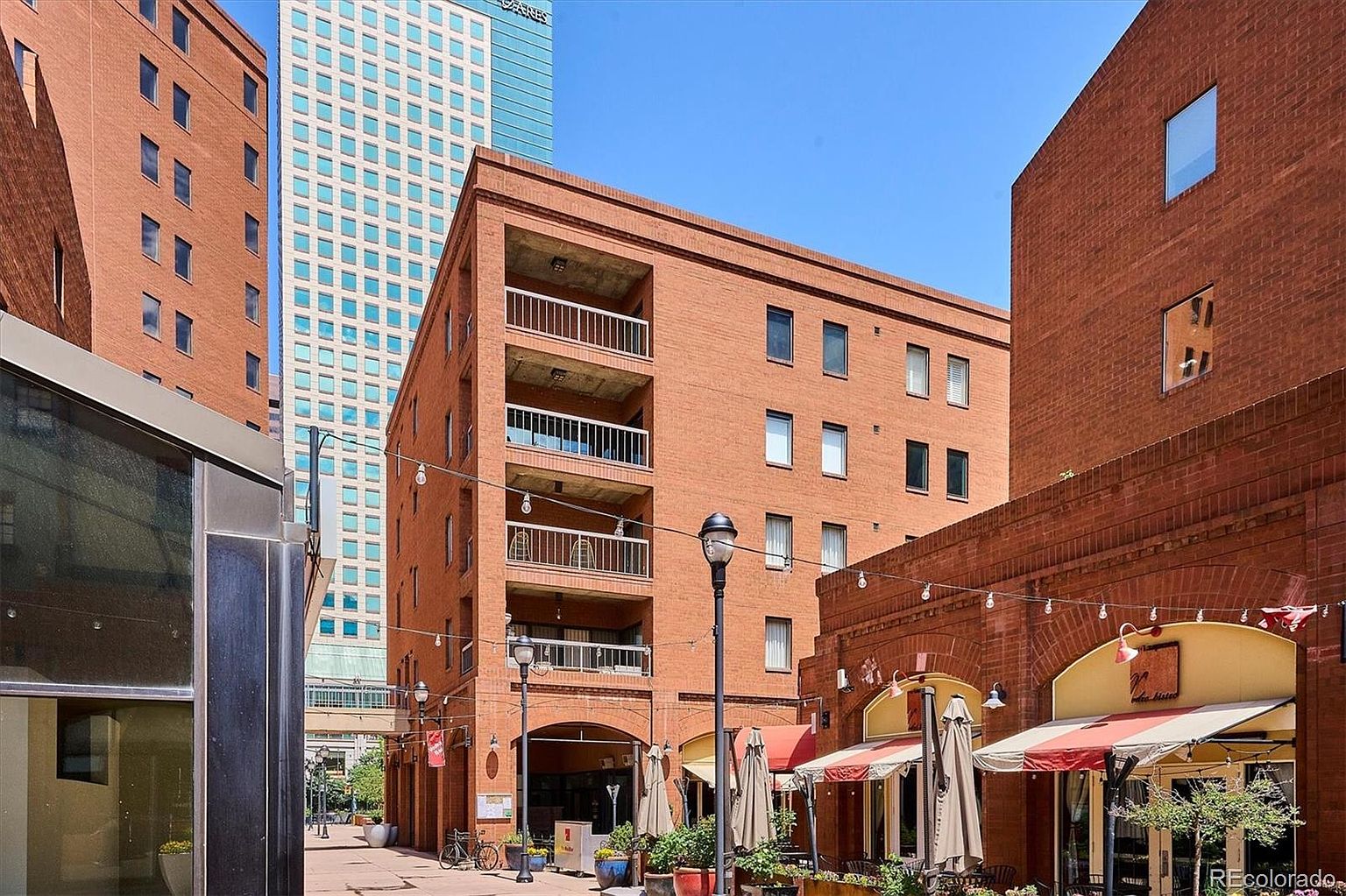 This image showcases the front exterior of a brick building with multiple balconies, situated in a vibrant urban setting. The building features a classic brick facade, complemented by white balcony railings and numerous windows. Street-level businesses with awnings and outdoor seating add to the lively atmosphere, suggesting a desirable location with convenient access to amenities.