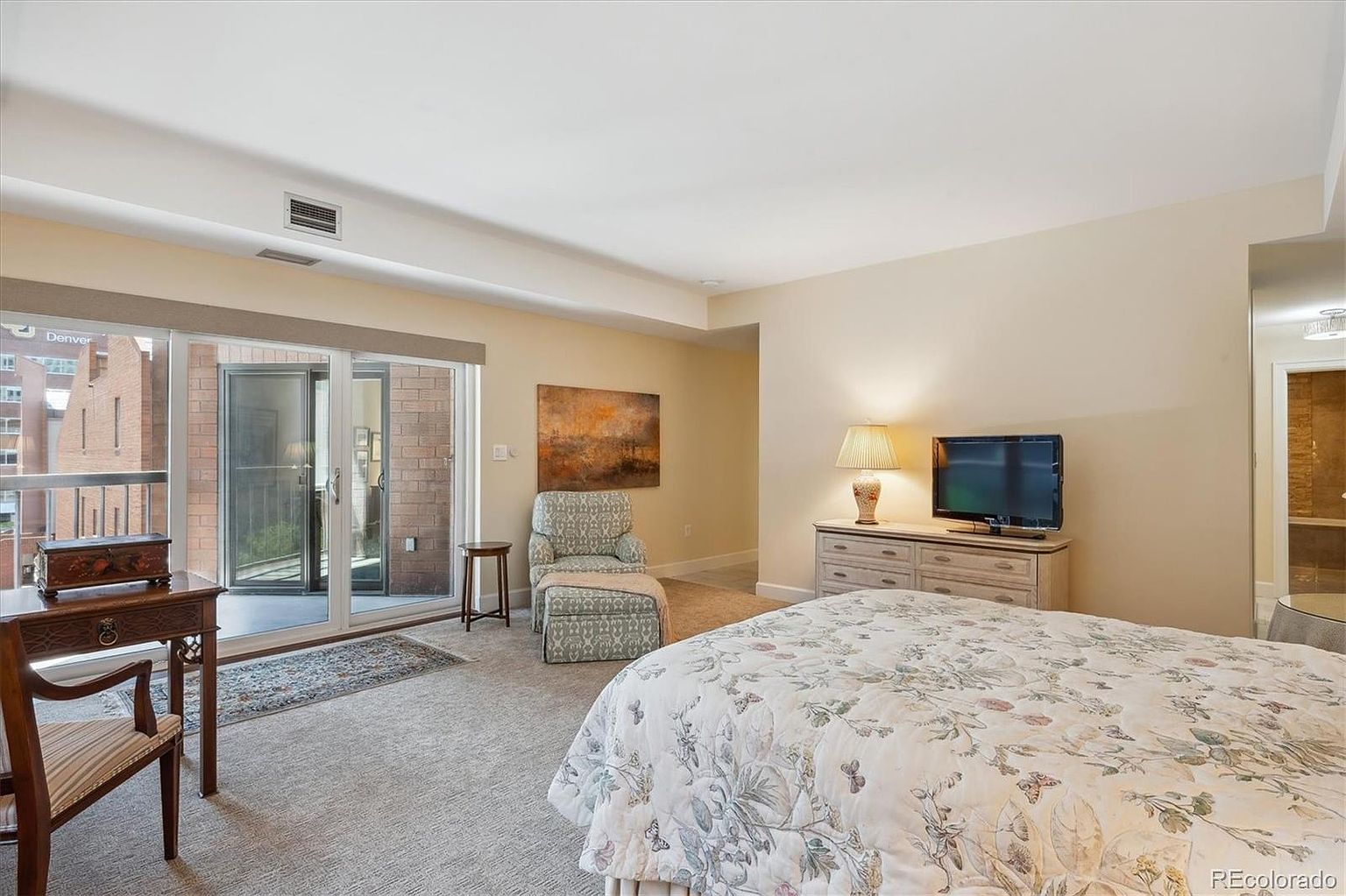 This is a primary bedroom featuring a sliding glass door to a balcony, a patterned armchair with ottoman, and a dresser with a television. The room is carpeted and has neutral-colored walls, creating a calm and inviting atmosphere. The bed is covered with a floral patterned comforter.