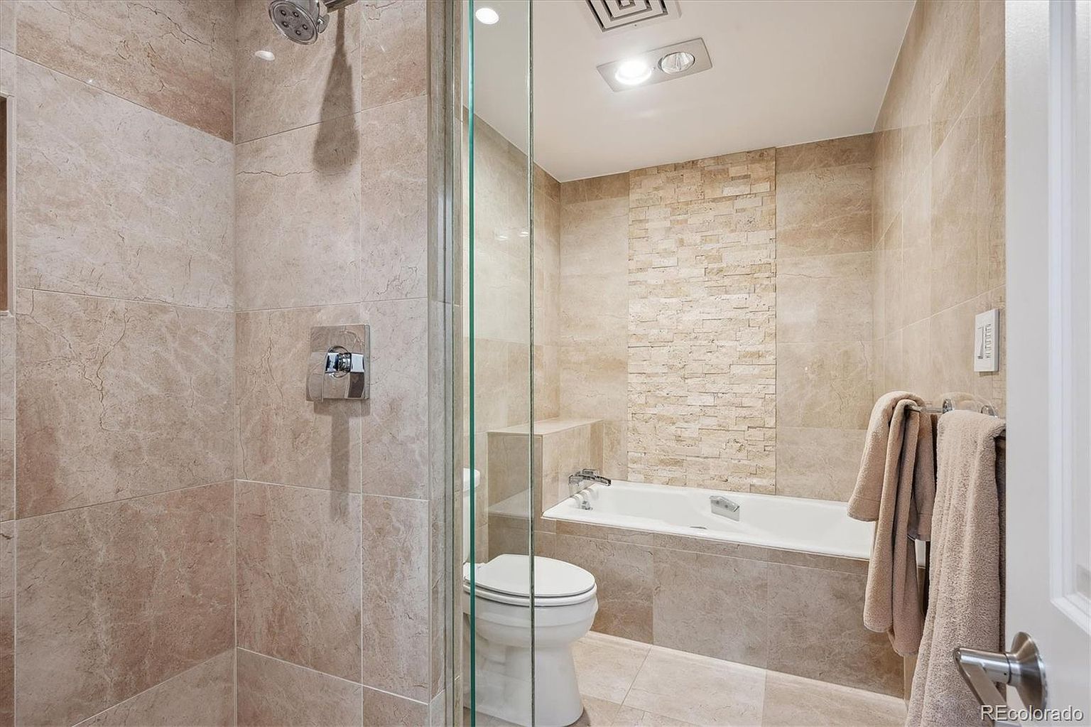 This is a primary bathroom featuring a walk-in shower with glass enclosure and a separate bathtub. The walls and floors are covered in light beige marble tiles, creating a luxurious and cohesive look. A modern toilet is visible, and the overall impression is clean and spa-like.