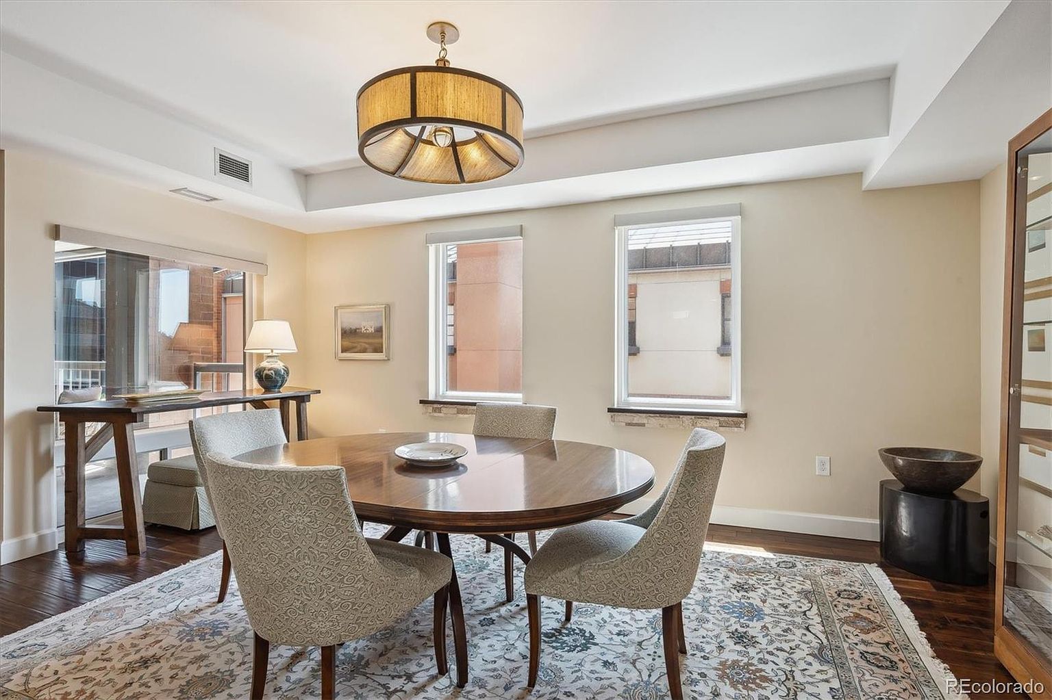 This is an interior shot of a dining room featuring a round wooden table with four upholstered chairs. A drum-style chandelier hangs above the table, and natural light streams in through two windows. The room is decorated in neutral tones with a patterned area rug, creating a warm and inviting atmosphere.