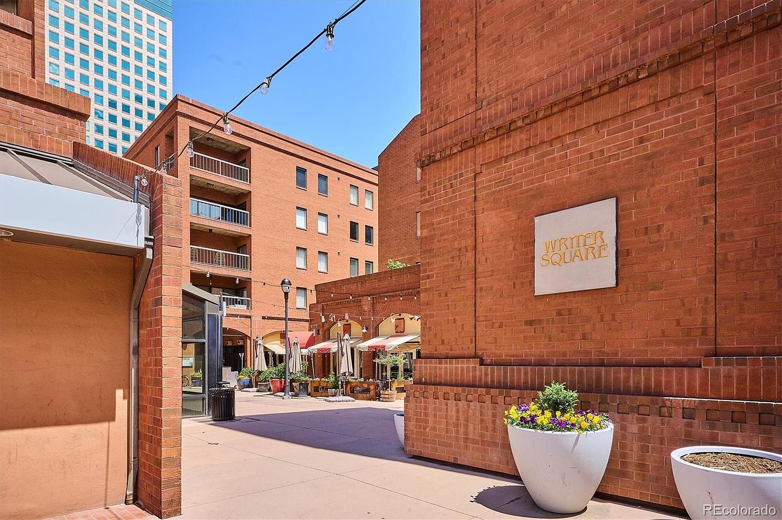 This exterior shot showcases a charming brick-paved square, possibly a community amenity. The scene features well-maintained brick buildings, planters with colorful flowers, and outdoor seating areas under awnings, creating an inviting and pedestrian-friendly atmosphere. A sign reading "Writer Square" is visible on one of the brick walls.