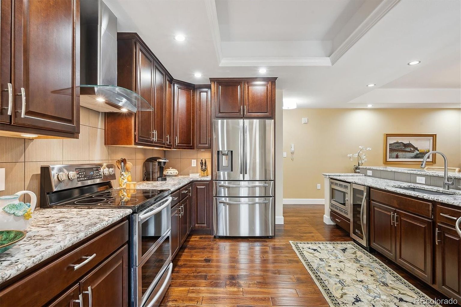 This is a well-lit kitchen featuring dark wood cabinetry, granite countertops, and stainless steel appliances, including a refrigerator, oven, and range hood. The hardwood flooring adds warmth to the space, and the overall design is elegant and functional. The kitchen also features a wine cooler and a small rug.