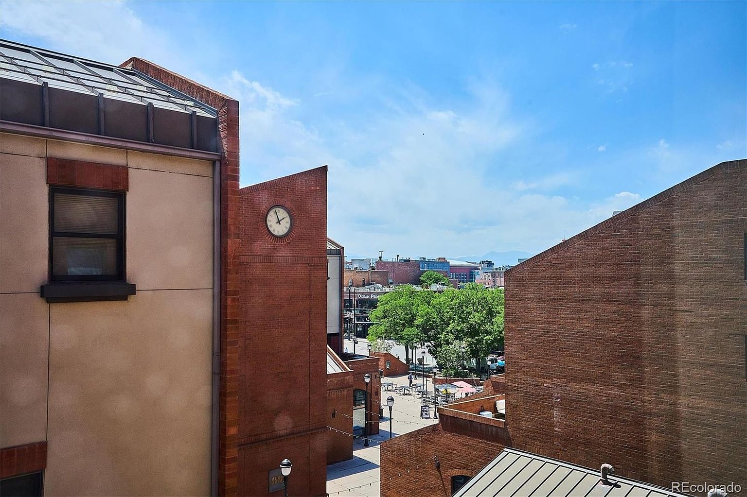 This exterior shot showcases a building's side view, emphasizing its brick facade and architectural details like a prominent clock. The image captures a partial view of a courtyard or plaza below, suggesting a vibrant urban environment. The clear blue sky and partial view of distant buildings add depth and context to the property's location.