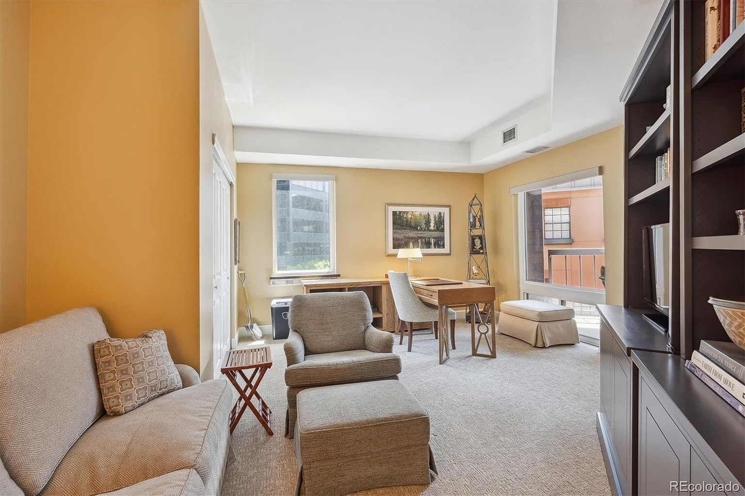 This is an interior shot of a home office or study. The room features a desk with a chair, a comfortable armchair with an ottoman, and a sofa with a decorative pillow. A built-in bookcase is visible on the right side of the image. The room is well-lit with natural light coming through the windows, and the overall style is traditional and cozy.