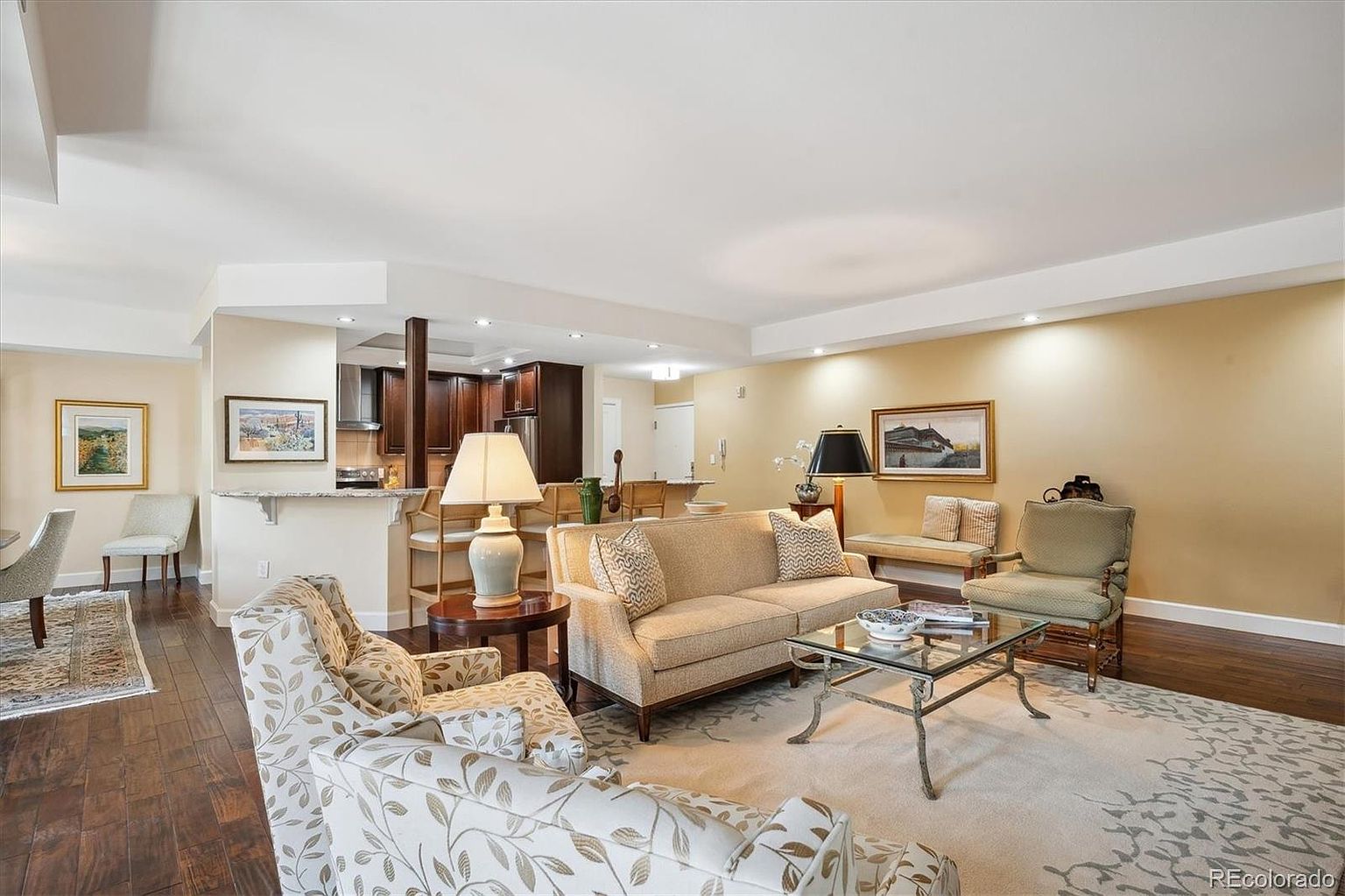 This is an interior shot of a well-decorated living room. The room features a neutral color palette with hardwood floors, a patterned area rug, and comfortable seating including a sofa and armchairs. A glass-topped coffee table sits in the center, and artwork adorns the walls, creating a warm and inviting atmosphere.