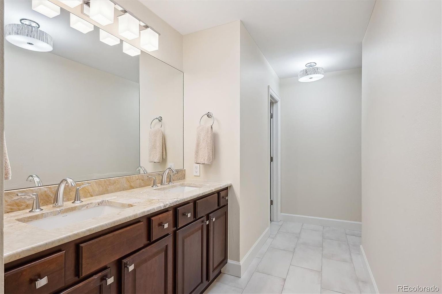 This is a well-lit primary bathroom featuring a double vanity with a light-colored countertop and dark wood cabinetry. A large mirror spans the length of the vanity, reflecting the overhead lighting. The bathroom extends into a hallway with tiled flooring and a doorway, creating a sense of spaciousness.