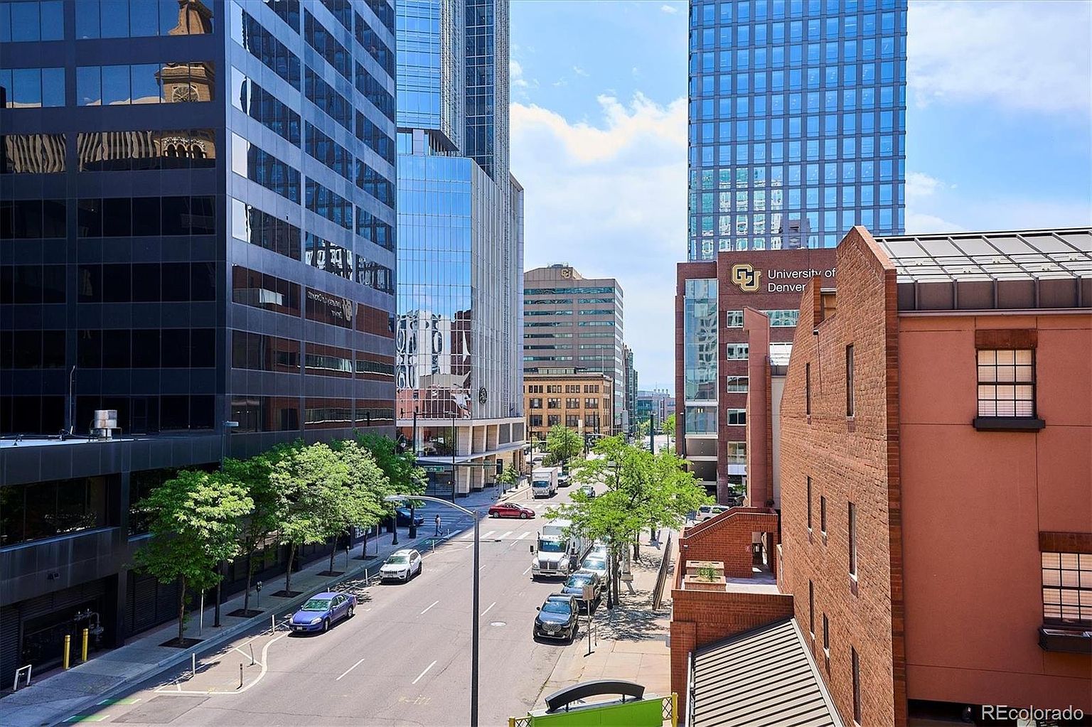 This image showcases a vibrant street view in a bustling urban setting. Tall buildings with reflective glass facades line the street, creating a modern and dynamic atmosphere. The street is active with cars and trees, adding a touch of nature to the cityscape. The University of Denver building is visible, adding an educational element to the scene.