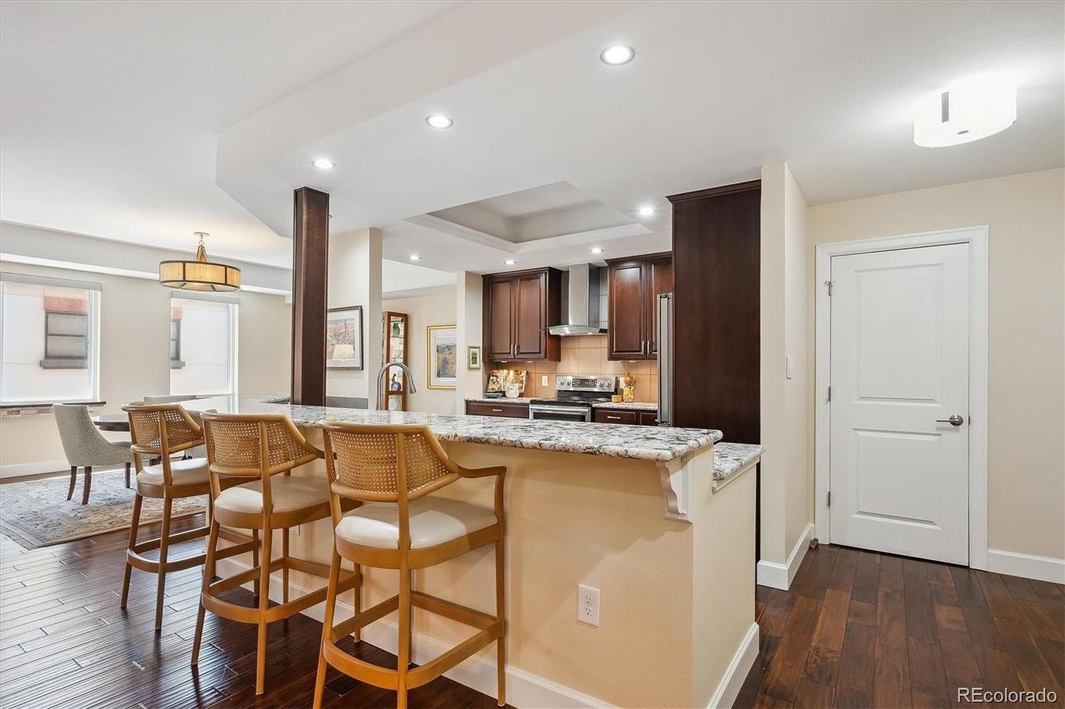 This interior shot showcases a well-lit kitchen with dark wood cabinetry and granite countertops. A breakfast bar with three stools provides seating, and the hardwood flooring adds warmth to the space. The kitchen seamlessly blends into an adjacent dining area, creating an open and inviting atmosphere.