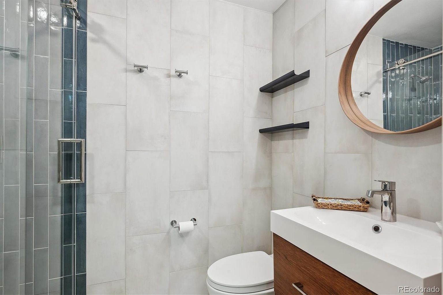 This is a well-lit bathroom featuring a modern design with light gray tiled walls and a wood-toned vanity. A round mirror hangs above the sink, reflecting the shower area with blue accent tiles. The space is clean and minimalist, creating a serene and functional atmosphere.