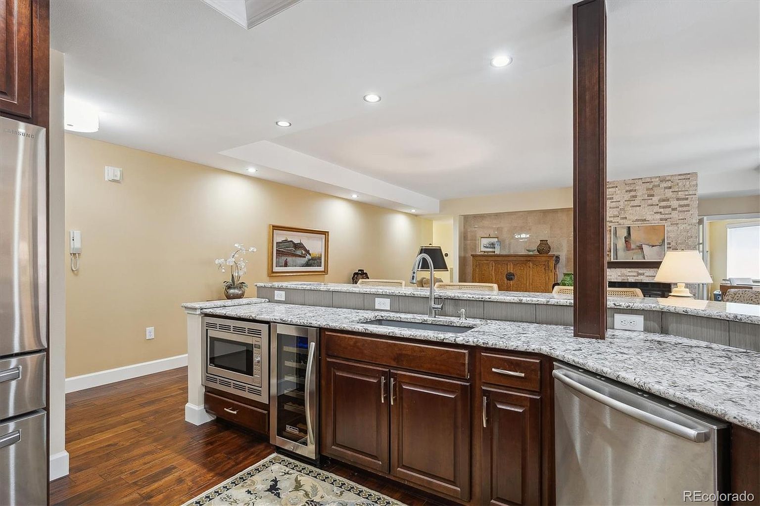 This interior shot showcases a well-appointed kitchen area with dark wood cabinetry, granite countertops, and stainless steel appliances, including a microwave, wine cooler, and dishwasher. The kitchen features a sink with a modern faucet and is adjacent to a living space, separated by a half-wall, creating an open and inviting atmosphere. The flooring is hardwood, and the overall impression is one of warmth and functionality.