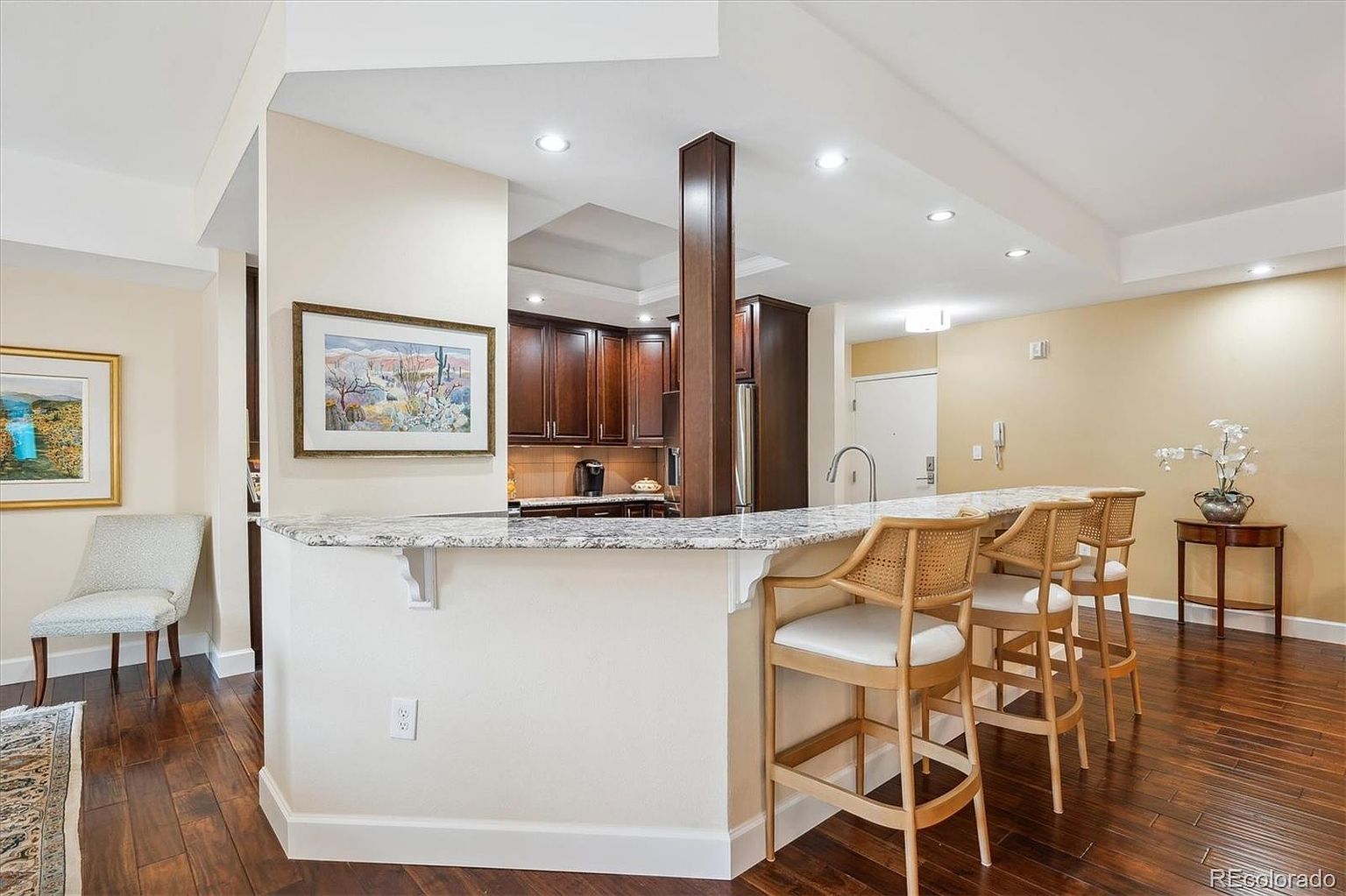 This interior shot showcases a well-appointed kitchen with dark wood cabinetry and a granite countertop breakfast bar. Three bar stools with light wood frames and white cushions line the bar, and the hardwood flooring adds warmth to the space. The kitchen is open to a living area, creating a welcoming and functional layout.