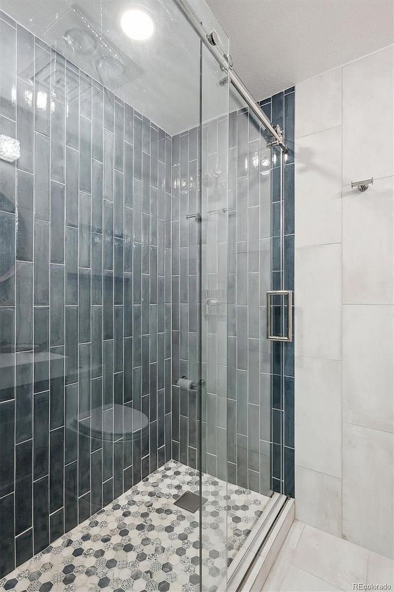 This is a well-lit primary bathroom featuring a modern shower with a sliding glass door. The shower walls are tiled with vertical, gray-blue rectangular tiles, while the floor is covered in a mosaic of hexagonal tiles. A toilet is partially visible behind the shower glass, and the overall impression is clean and contemporary.