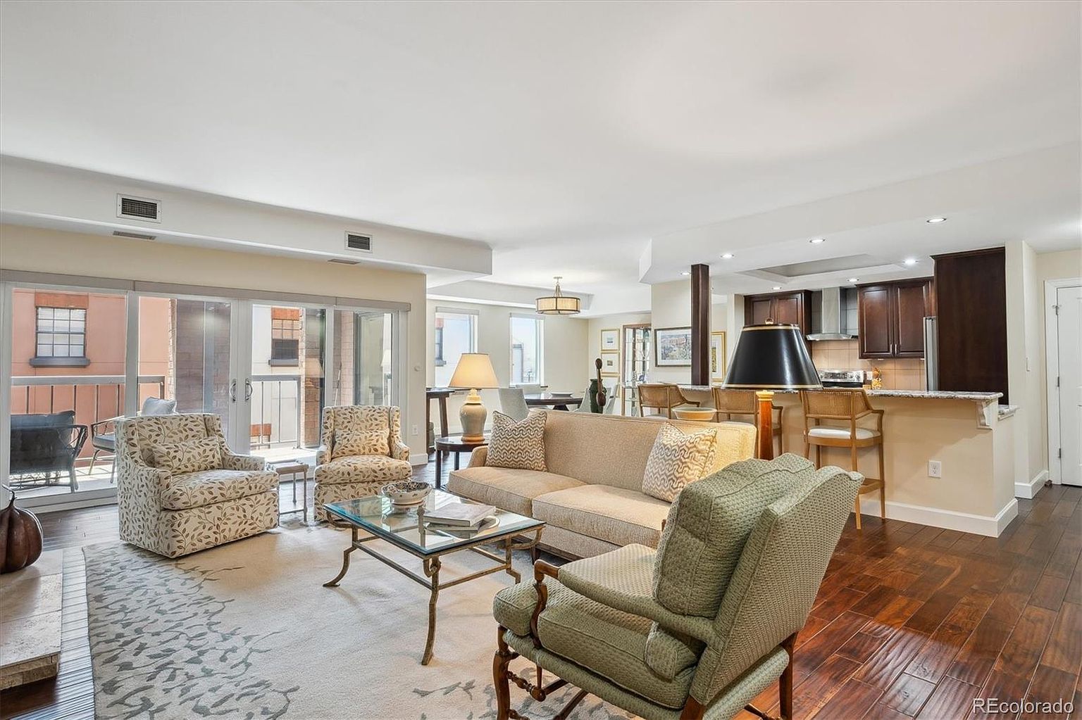 This is an interior shot of a well-lit living room that flows into a kitchen area. The living room features a neutral color palette with patterned armchairs, a beige sofa, and a glass-topped coffee table on a patterned rug. The kitchen has dark wood cabinetry and a breakfast bar with stools, creating an open and inviting space.