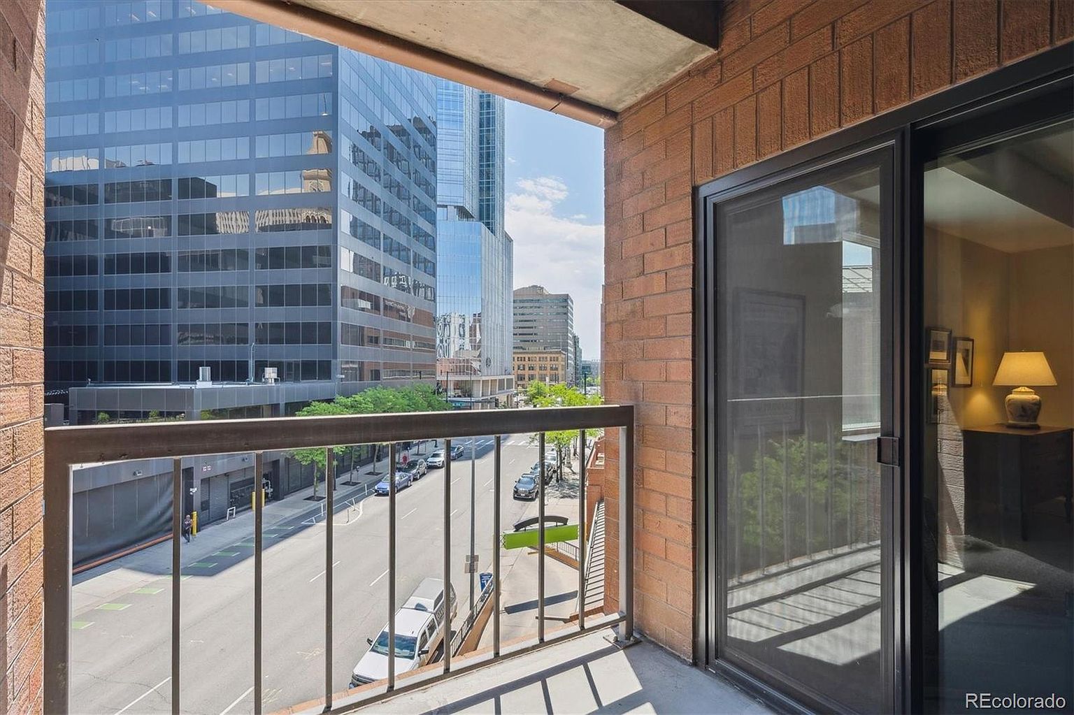 This image showcases a balcony with a view of a city street and surrounding buildings. The balcony features a metal railing and a sliding glass door leading to an interior space. The view includes modern skyscrapers and street-level activity, creating an urban ambiance.