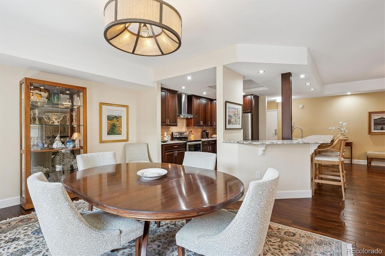 This interior shot showcases a dining room with a round wooden table surrounded by upholstered chairs. A glass-front cabinet displays decorative items, and a modern chandelier hangs above the table. The dining area transitions into a kitchen space, creating an open and inviting atmosphere.