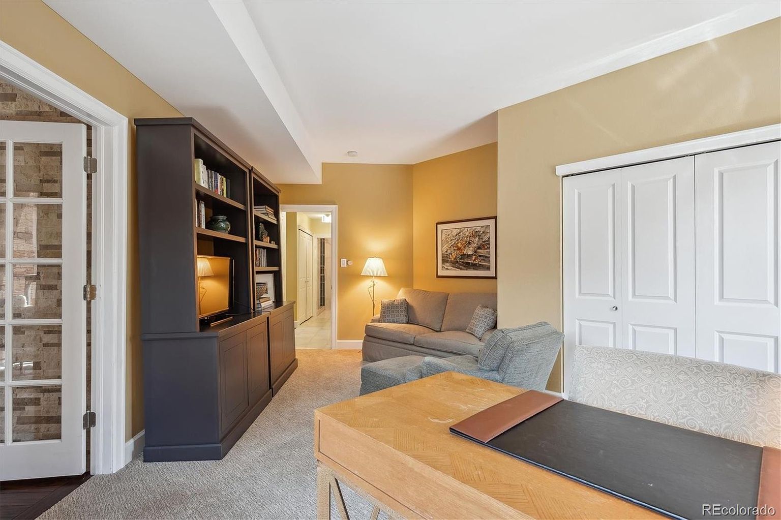 This is an interior shot of a home office or study. The room features a built-in bookcase with a television, a desk with a leather writing surface, and a seating area with a sofa and armchair. The walls are painted in a warm, neutral tone, and the flooring is carpeted, creating a cozy and functional workspace.