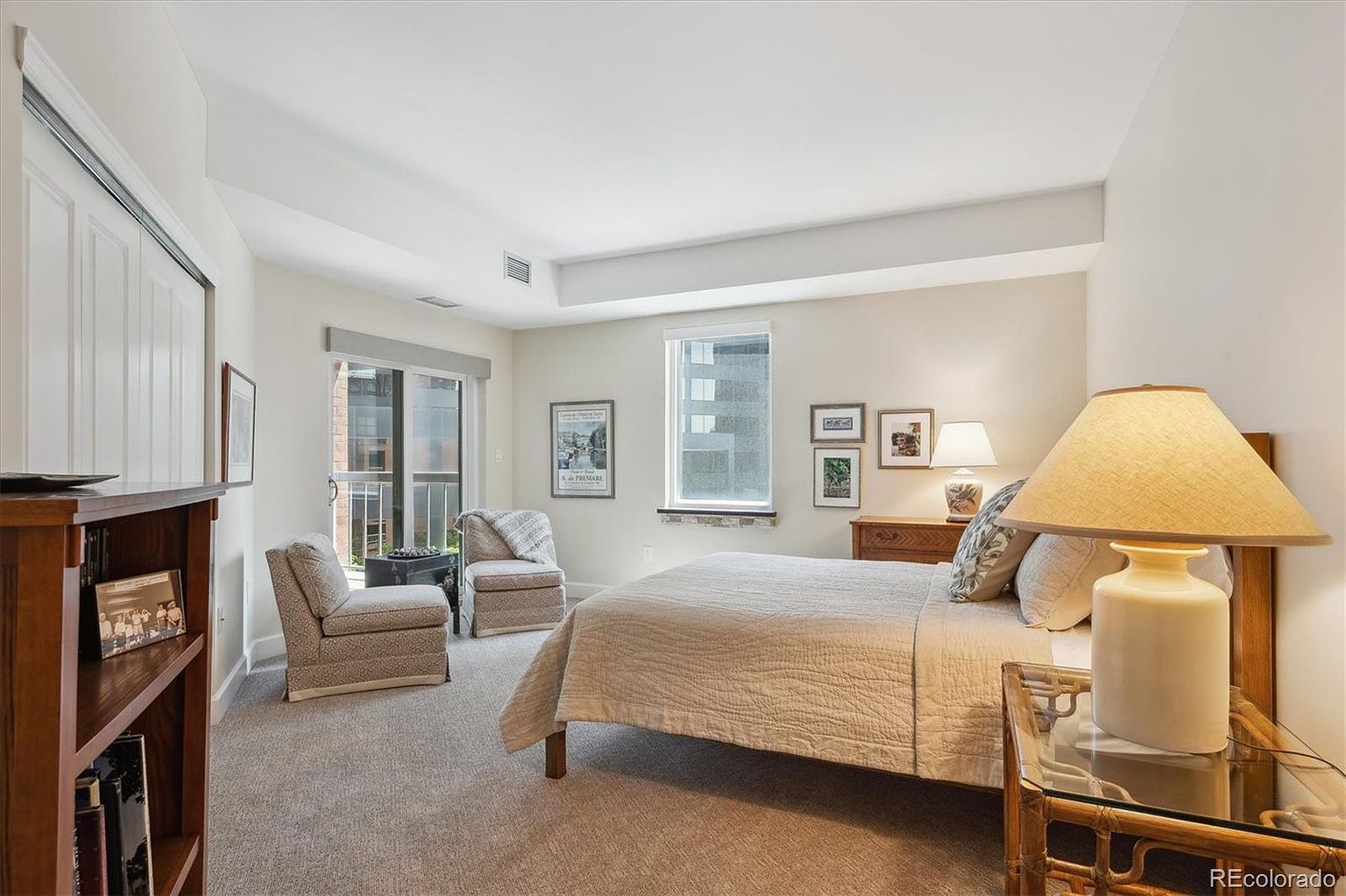 This is a well-lit primary bedroom featuring a neutral color palette. The room includes a bed with a light-colored quilt, two armchairs near a balcony door, and a wooden dresser. A side table with a lamp sits next to the bed, and the room is carpeted, creating a cozy and inviting atmosphere.