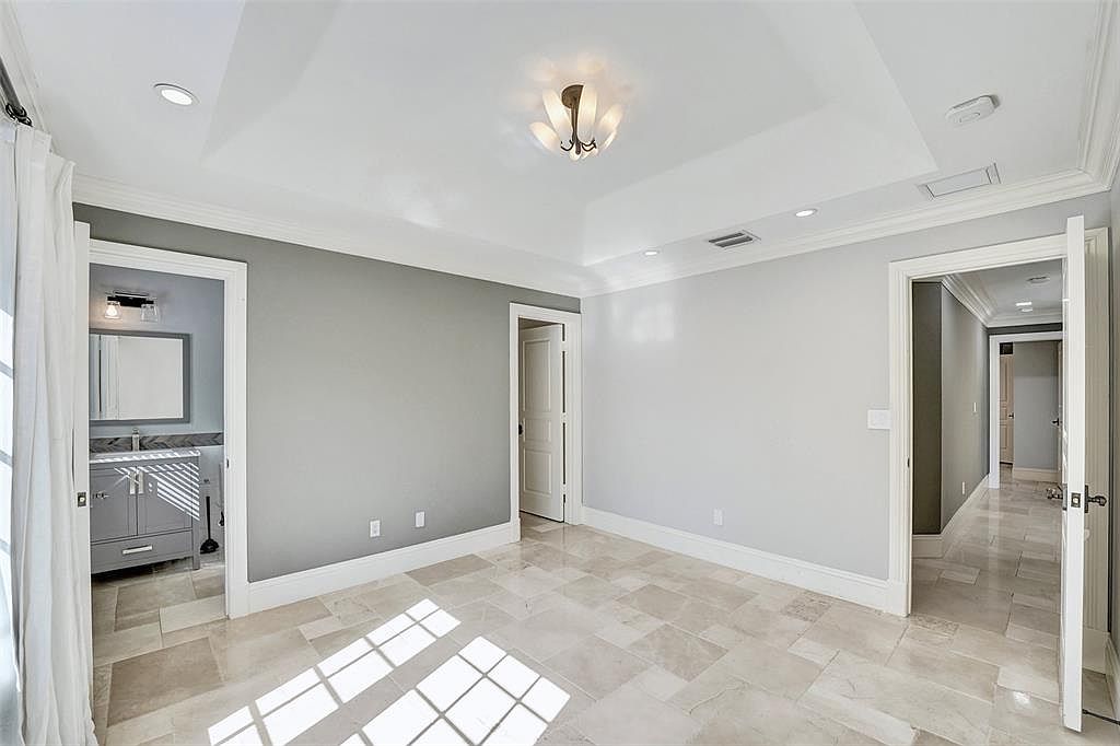 This is a primary bedroom featuring a tray ceiling with a decorative light fixture and recessed lighting. The walls are painted in a neutral gray tone, and the flooring is tiled with large, light-colored tiles. There are doorways leading to a bathroom and hallway, enhancing the sense of spaciousness and connectivity.