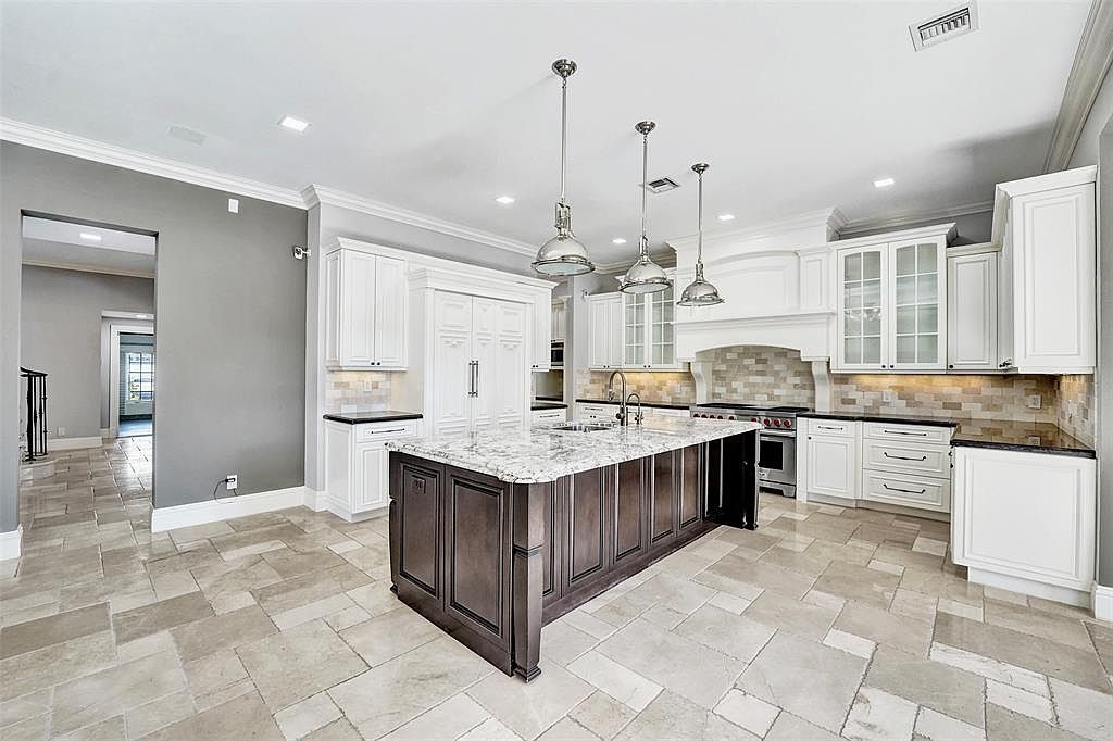 This is a spacious kitchen featuring white cabinetry, dark countertops, and a large center island with a light-colored countertop. Three pendant lights hang above the island, and stainless steel appliances are visible. The flooring is a light-colored tile, and the overall impression is one of luxury and functionality.