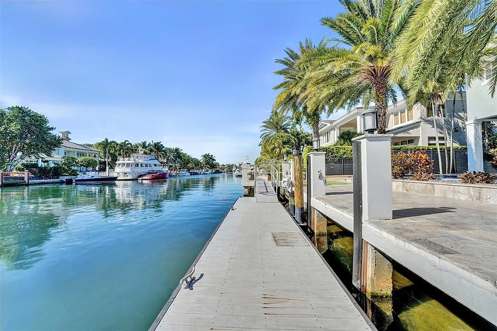 This image showcases the side of a waterfront property, emphasizing the dock and canal. The dock extends into the water, providing boat access, while palm trees and modern architecture add to the luxurious feel. The clear blue sky and calm water create a serene and inviting atmosphere.