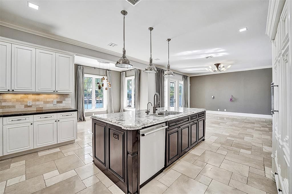 This is a bright and spacious kitchen featuring a large center island with a granite countertop and dark wood cabinetry. The kitchen also includes white cabinets along the walls, stainless steel appliances, and a tile backsplash. Large windows provide natural light and views of the outdoors, enhancing the open and airy feel of the space.