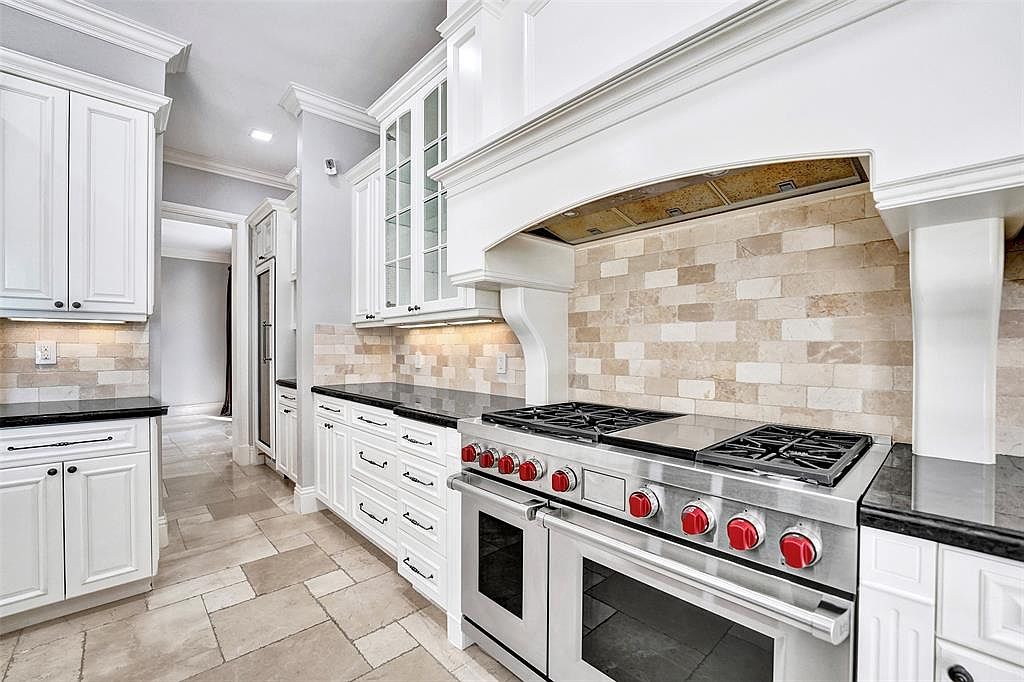 This is a well-lit kitchen featuring white cabinetry, black countertops, and stainless steel appliances, including a professional-grade range with red knobs. The backsplash is a neutral-toned brick pattern, and the flooring is a light-colored tile. The kitchen has a clean and elegant design, with a focus on functionality and style.