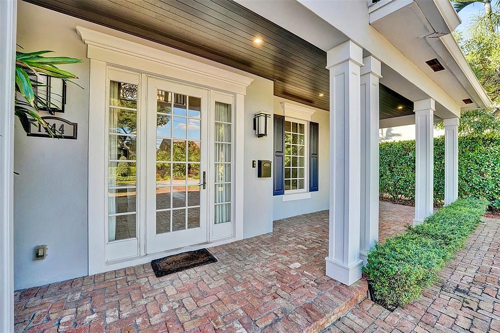 This image showcases the entryway of a charming home, featuring a brick walkway leading to a set of white French doors. The entrance is framed by white columns and trim, with a dark wood ceiling overhead. A neatly trimmed hedge lines the walkway, adding to the curb appeal and creating a welcoming atmosphere.