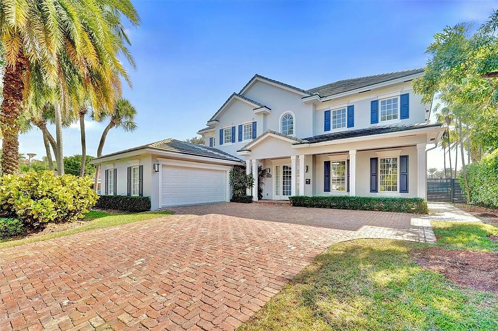 This is a front exterior view of a two-story house with a light gray facade and dark blue shutters. The house features a brick driveway, a two-car garage on the left, and well-maintained landscaping. The overall impression is one of a well-kept, upscale home in a sunny location, with palm trees adding to the tropical feel.