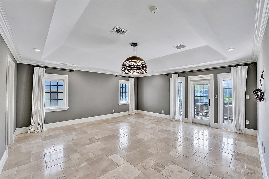This is a spacious living room featuring neutral gray walls, a light-colored tile floor, and a modern pendant light fixture. The room has multiple windows and glass doors leading to an outdoor area, providing ample natural light. The overall impression is clean, bright, and inviting, suitable for a comfortable living space.