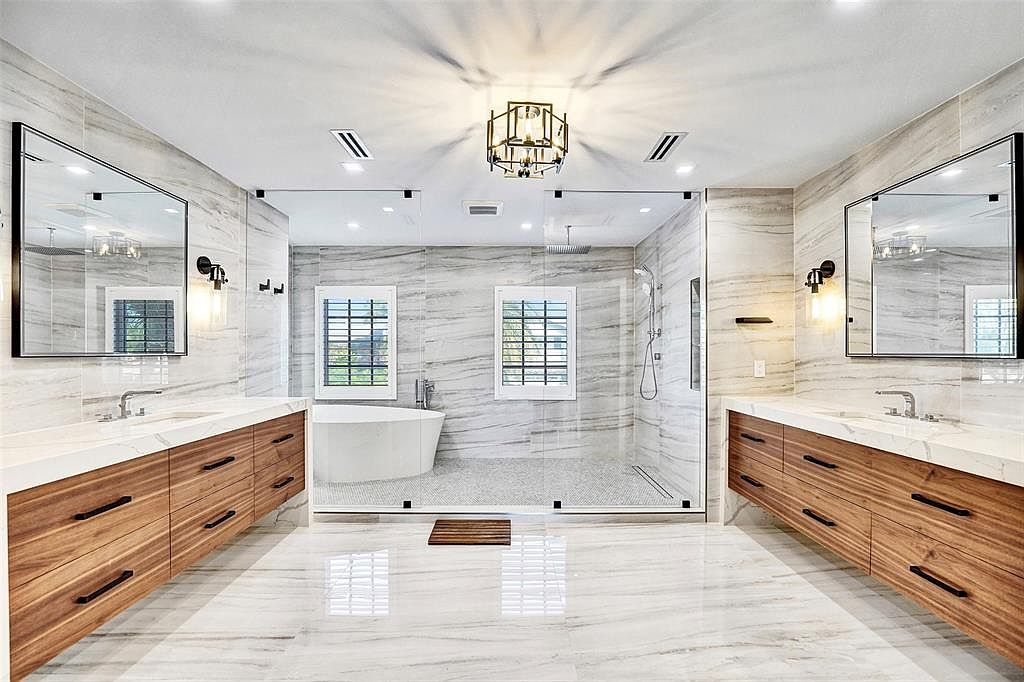 This is a luxurious primary bathroom featuring a spacious walk-in shower enclosed with glass, a freestanding bathtub, and dual vanities with modern wood cabinetry and marble countertops. The walls and floors are covered in marble tiles, and the room is illuminated by a stylish chandelier, creating a bright and elegant atmosphere. The perspective is a wide shot, showcasing the entire layout of the bathroom.