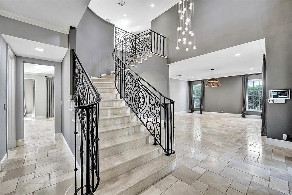This interior shot showcases a grand foyer with a winding staircase featuring ornate black iron railings and light-colored stone steps. The walls are painted in a sophisticated gray, complemented by light-colored tile flooring. A modern chandelier hangs in the background, adding a touch of elegance to the space, while an open doorway leads to a living area with large windows.